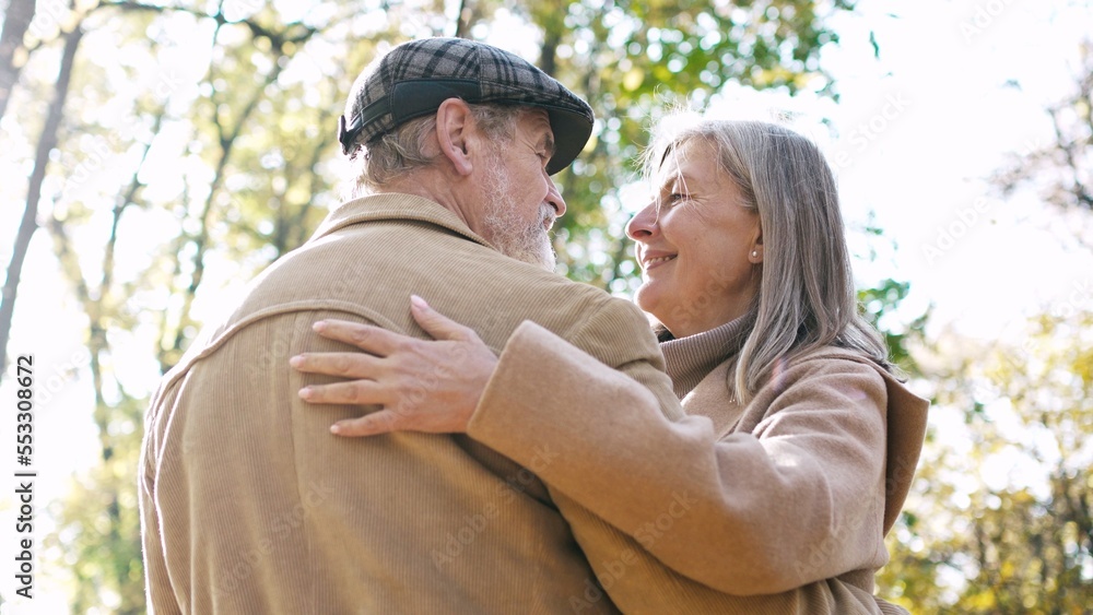 Beautiful cute mature grey-haired woman hugging elegant bearded beautiful man standing in autumn park wearing coats. Lovely couple spending time on fresh air together looking at each other. Beautiful cute mature grey-haired woman hugging elegant bearded beautiful man standing in autumn park wearing coats. Lovely couple spending time on fresh air together looking at each other.