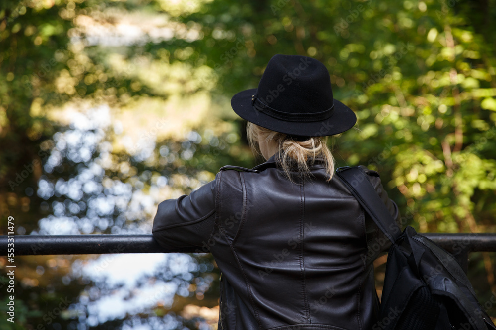 young Caucasian woman in a leather jacket, hat and with a backpack stands on the bridge and looks at the river. selective focus. High quality photo