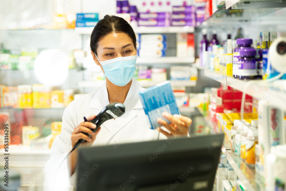 Asian female pharmacist in face mask standing at counter and using barcode scanner to sell pharmaceutical package.