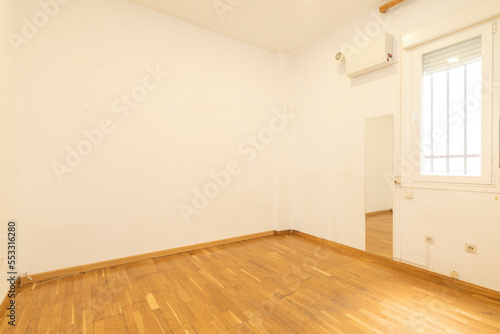 An empty room with a frameless mirror on the wall, worn wooden floors with scratch marks and a barred window