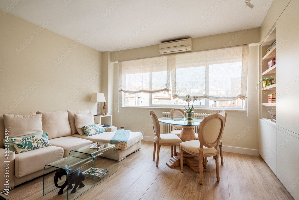 Fototapeta premium Living room decorated in a contemporary style with a round glass table, wooden chairs upholstered in plain fabric and a three-seater fabric sofa with a chaise longue and glass coffee tables