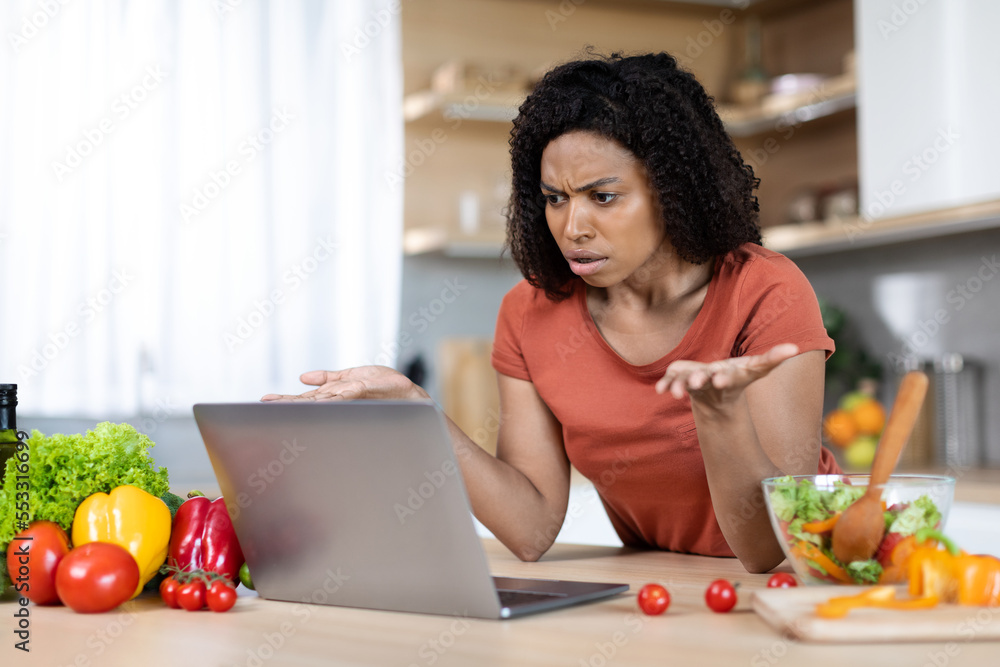 Sad angry young black lady gesturing with hands at computer webcam in kitchen interior