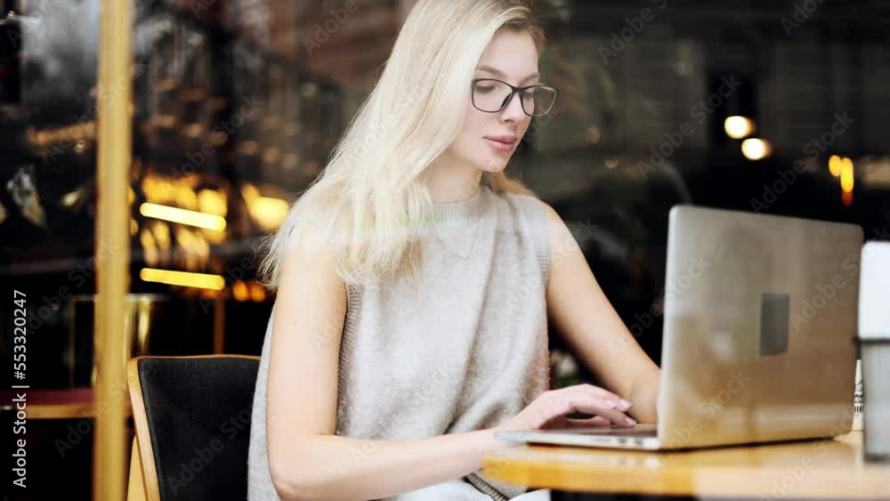 View through the window of charming young woman have distance remote freelance work on computer while sitting in street coffee shop alone Blond busy lady with glasses browsing typing on laptop View through the window of charming young woman have distance remote freelance work on computer while sitting in street coffee shop alone Blond busy lady with glasses browsing typing on laptop