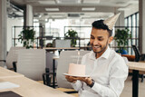 Happy Hindu businessman holding cake with lightened candle in the office and looking at camera.