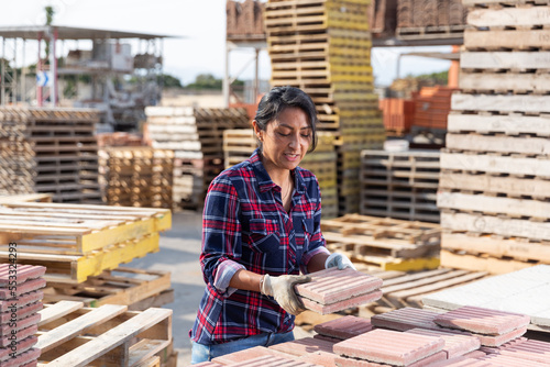 Concentrated hispanic woman worker in gloves stacking red paving slabs on pallet at hardware store warehouse