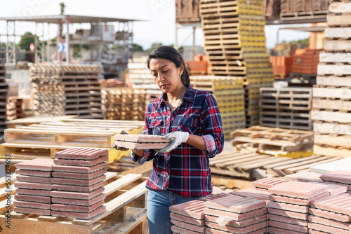 Concentrated hispanic woman worker in gloves stacking red paving slabs on pallet at hardware store warehouse