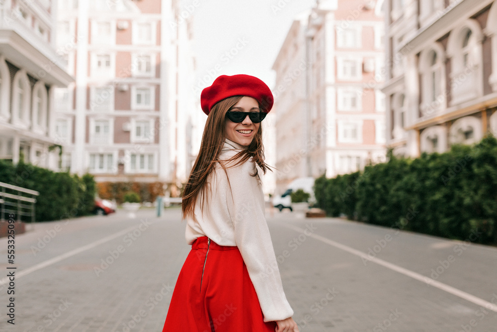 Attractive stylish woman in sunglasses and red hat on head, walking in park. Pretty woman with light brown hair smiles broadly teeth. Autumn fashion accessories in street fashion style.