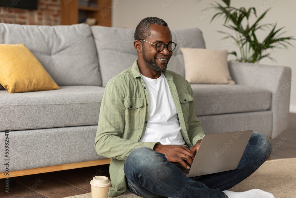 Smiling busy mature black guy in glasses typing on computer, watch video, sits on floor with cup of coffee