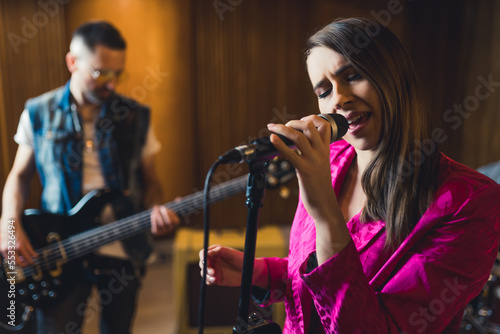medium closeup side view shot of a young singer woman wearing a pink blazer and a guitarist in the background. High quality photo