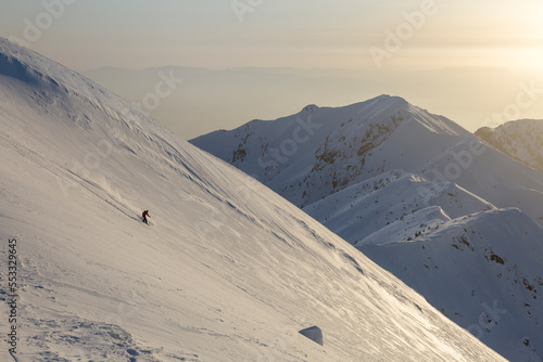 A freerider skier descends a wide slope against the background of mountain ranges and the sunset