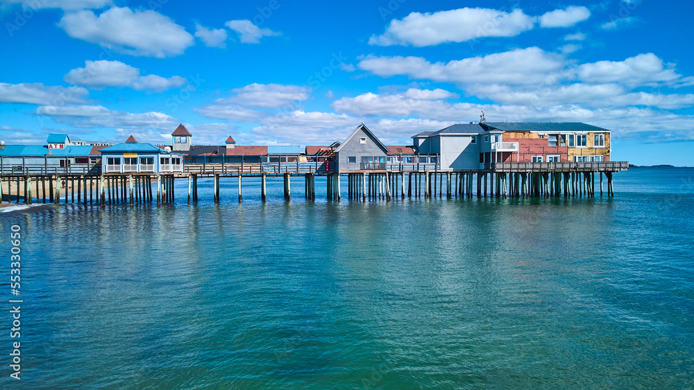 Aerial side profile of long old wood pier covered in shops in Maine with ocean and blue sky