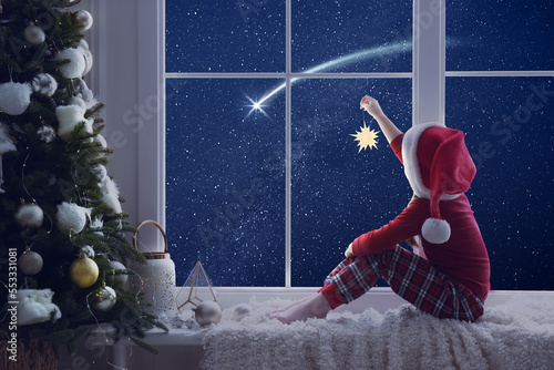 Cute little girl in Santa hat with Christmas ornament sitting on windowsill and looking at shooting star in beautiful night sky photo