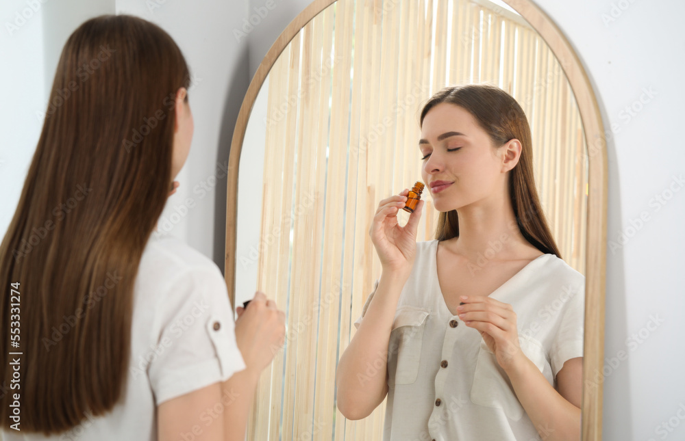 Young woman smelling essential oil near mirror indoors Young woman smelling essential oil near mirror indoors