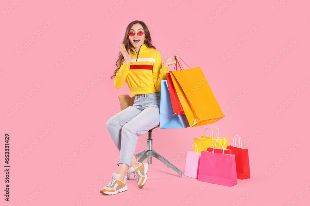 Emotional woman in stylish sunglasses holding many colorful shopping bags on armchair against pink background