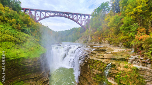 Canvas Print Steel maroon bridge over stunning raging waterfall cutting through layered cliff