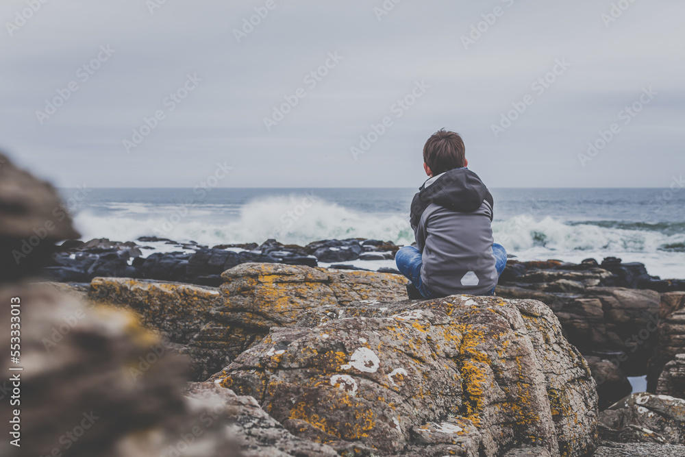 Young teen boy sitting on rocks and watching the sea, ocean, Atlantic Young teen boy sitting on rocks and watching the sea, ocean, Atlantic