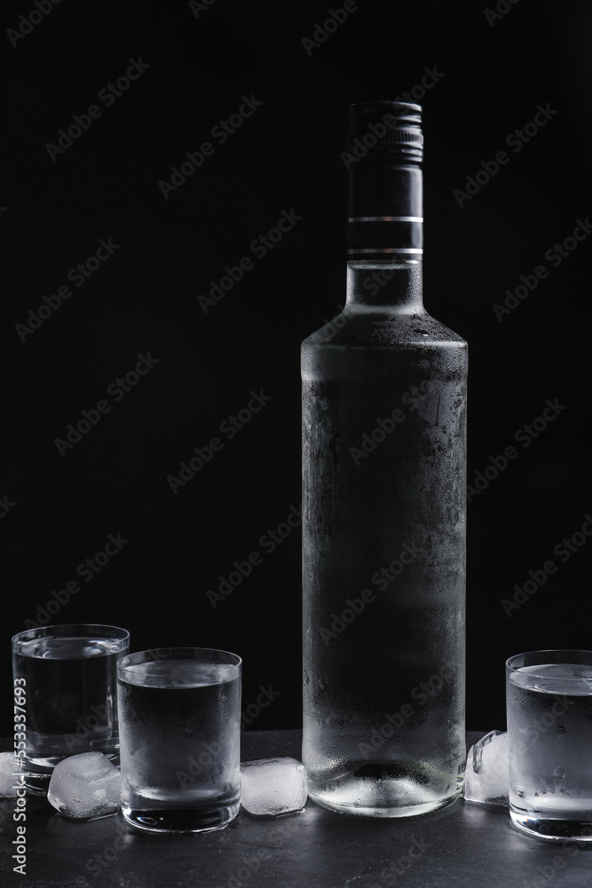 Bottle of vodka and shot glasses with ice on table against black background