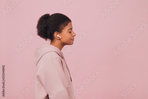 Profile close up portrait of black young woman with wireless headphones. Stylish girl with closed eyes in casual clothes listening music over pink backdrop