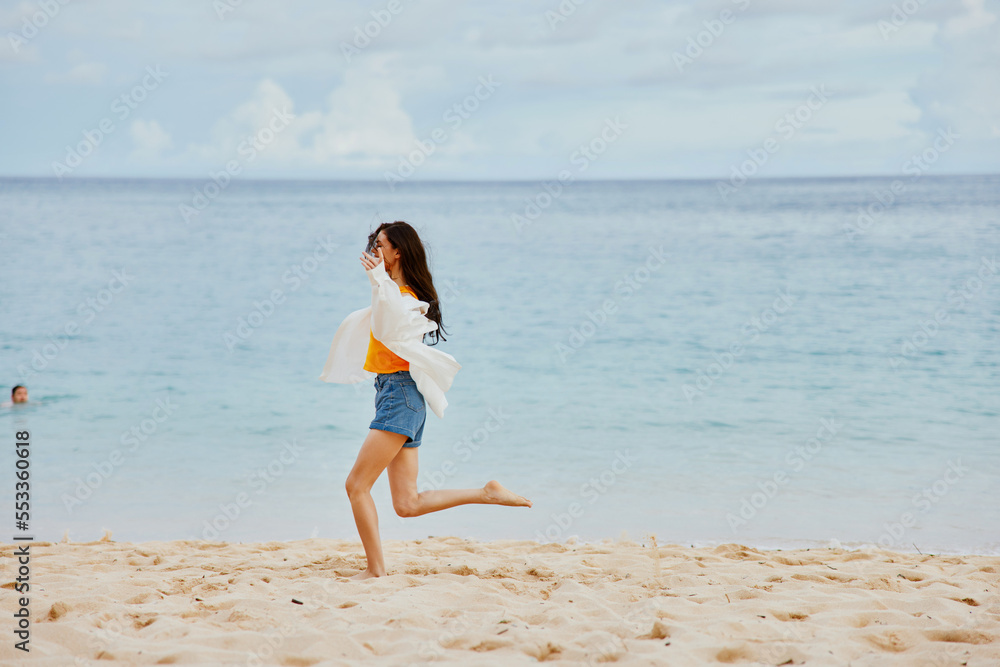 A woman runs along the beach in summer clothes on the sand in a yellow T-shirt and denim shorts white shirt flying hair ocean view