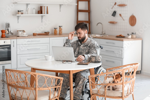 Young soldier in wheelchair using laptop at table in kitchen