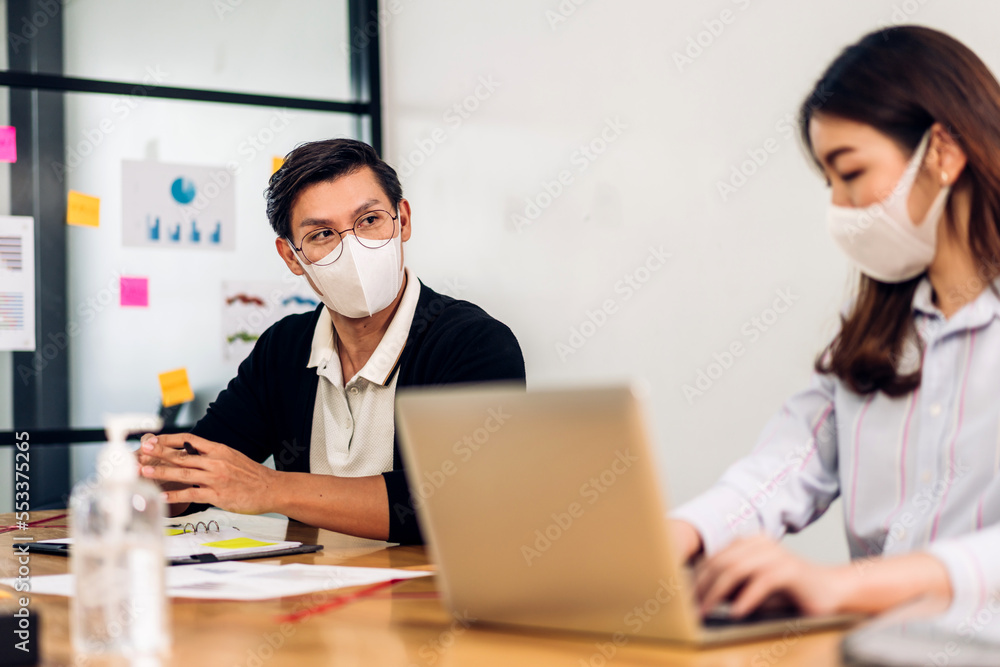 Asian business man and woman using laptop computer working and planning meeting in quarantine for coronavirus wearing protective mask with social distancing while sitting on office desk Asian business man and woman using laptop computer working and planning meeting in quarantine for coronavirus wearing protective mask with social distancing while sitting on office desk