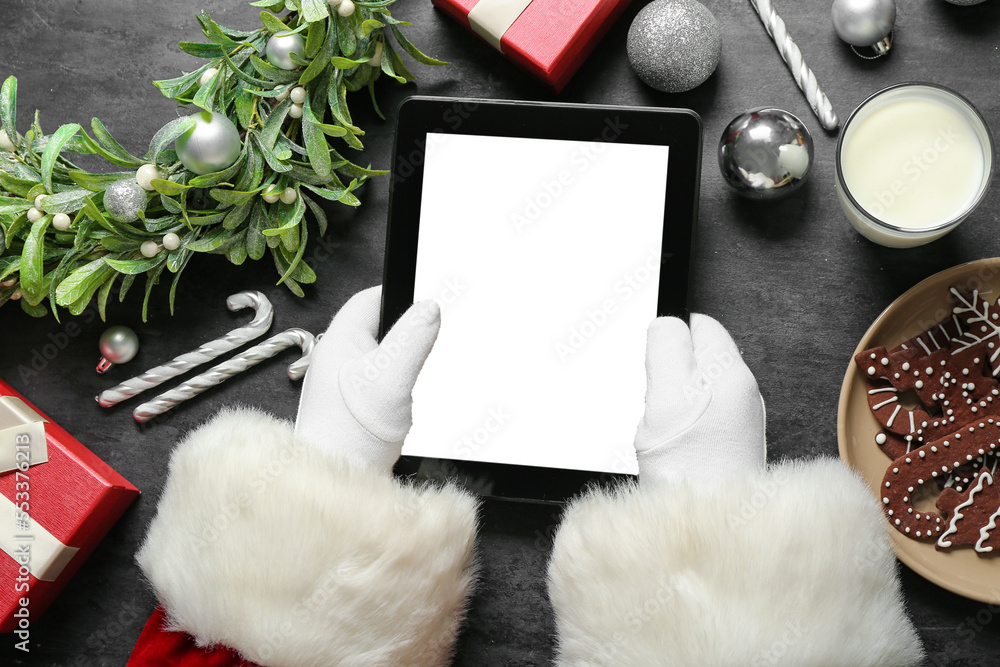 Santa Claus with tablet computer, glass of milk, cookies and Christmas decor on black background Santa Claus with tablet computer, glass of milk, cookies and Christmas decor on black background