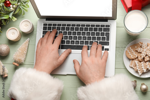 Santa Claus with glass of milk, cookies and Christmas decor using laptop on green wooden background