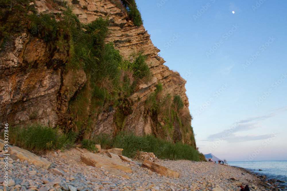 A steep section of a mountain with rock formations and trees on the seashore against the sky.