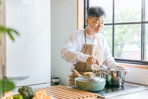 家のキッチンでフライパンを使って手料理を作るアジア人の高齢者男性（笑顔）
