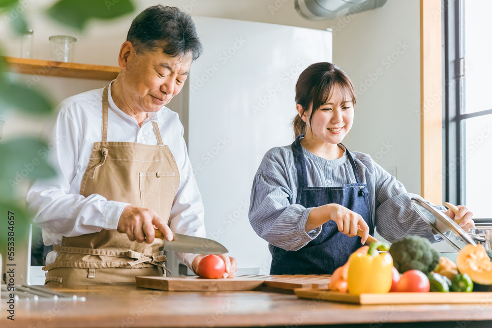 家のキッチンで一緒に料理・家事・自炊をする笑顔のアジア人高齢者男性と若い女性（家政婦・家事代行）
