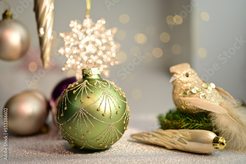 Decoration with a green Christmas ball with golden ornaments on a white tablewith a bird with sequins, a decorative stalactite, a snow star, Christmas balls and a blurred background with white lights photo
