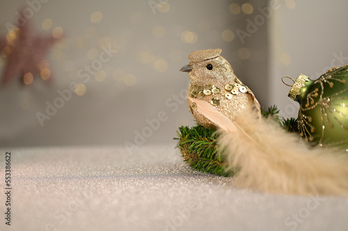 Christmas decoration bird with sequins and long tail feathers on an artificial fir branch on a white table with a Christmas ball and a blurred background with white lights