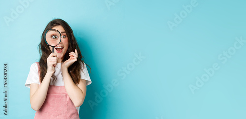 Happy young woman looking through magnifying glass with excited face, found or search something, standing over blue background
