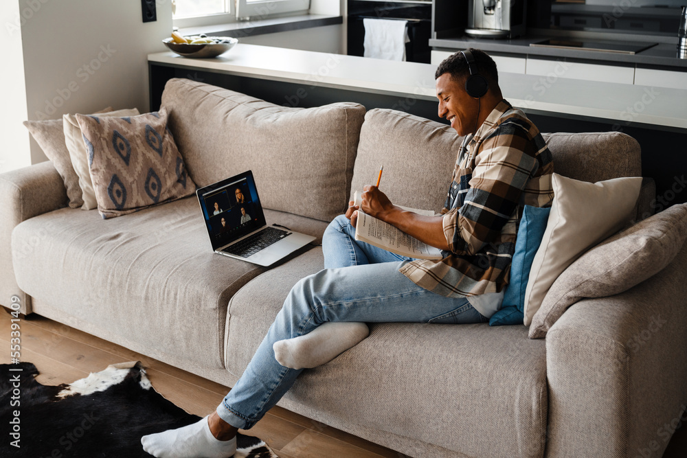 Obraz premium Afro man making online conference call on laptop and making notes while sitting on sofa