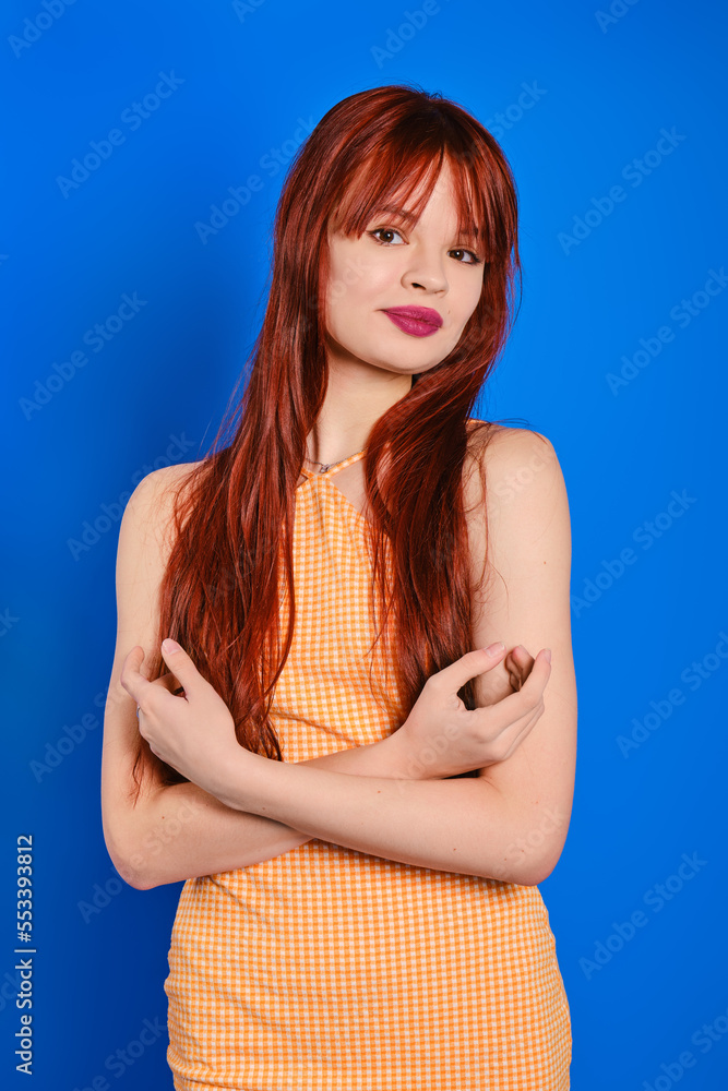 Slender young woman posing with crossed arms in peach spaghetti strap dress on blue background Slender young woman posing with crossed arms in peach spaghetti strap dress on blue background