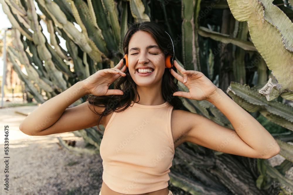 Positive young caucasian girl listening music with closed eyes stands near big cactuses. Brunette wears light top. Concept of devices.