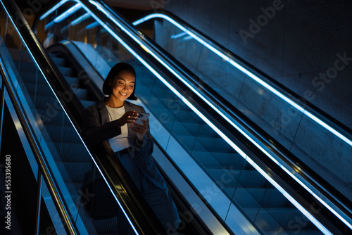 Young asian woman using mobile phone while standing on escalator indoors