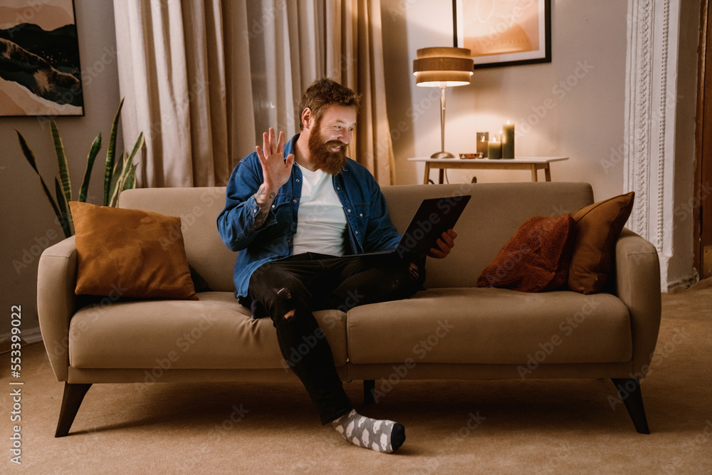 Smiling man having video call via laptop computer and waving while sitting on sofa