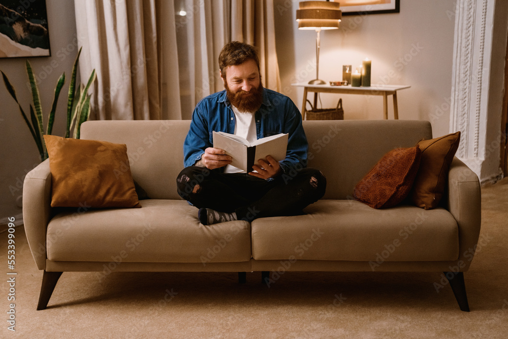 Cheerful bearded man reading book while sitting on sofa at home