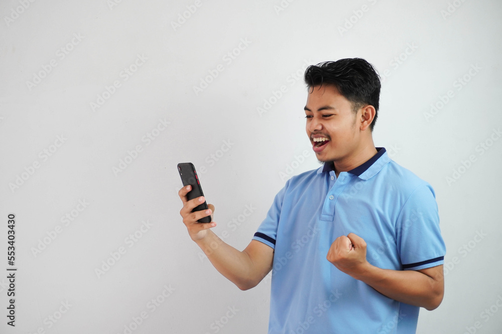 excited or happy portrait young asian man holding phone with clenched fists wearing blue polo t shirt isolated on white background excited or happy portrait young asian man holding phone with clenched fists wearing blue polo t shirt isolated on white background