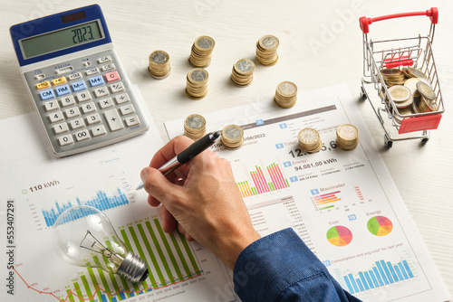 hand of a man with a pen on some electricity bills surrounded by coins a light bulb and a calculator