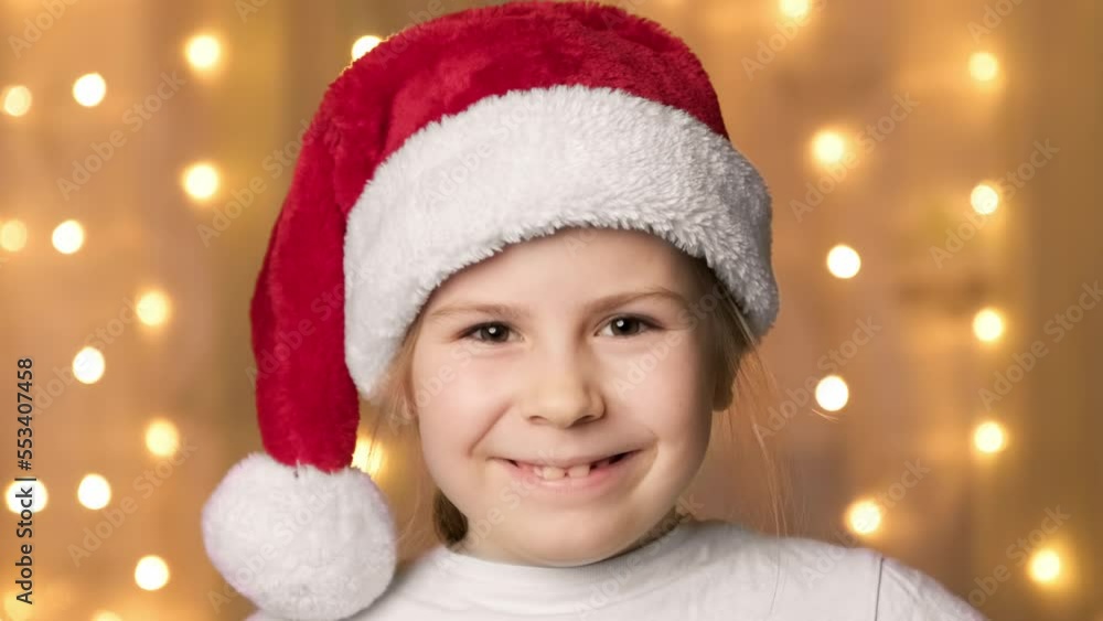 Christmas concept with little girl in christmas hat and garland on background. Adorable little girl with Santa hat. Happy child. Close-up in 4K, UHD Christmas concept with little girl in christmas hat and garland on background. Adorable little girl with Santa hat. Happy child. Close-up in 4K, UHD