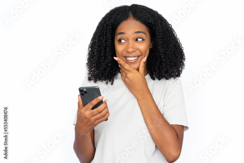 Excited African American woman with phone rubbing chin. Portrait of happy young female model with dark curly hair in white T-shirt looking away smiling, texting on phone. Modern technology concept