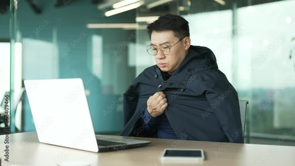 Cold asian businessman IT programmer suffers and trembles warms hands covering jacket try to work on laptop computer at desk in the modern office with problem of heating system indoors Low temperature Cold asian businessman IT programmer suffers and trembles warms hands covering jacket try to work on laptop computer at desk in the modern office with problem of heating system indoors Low temperature