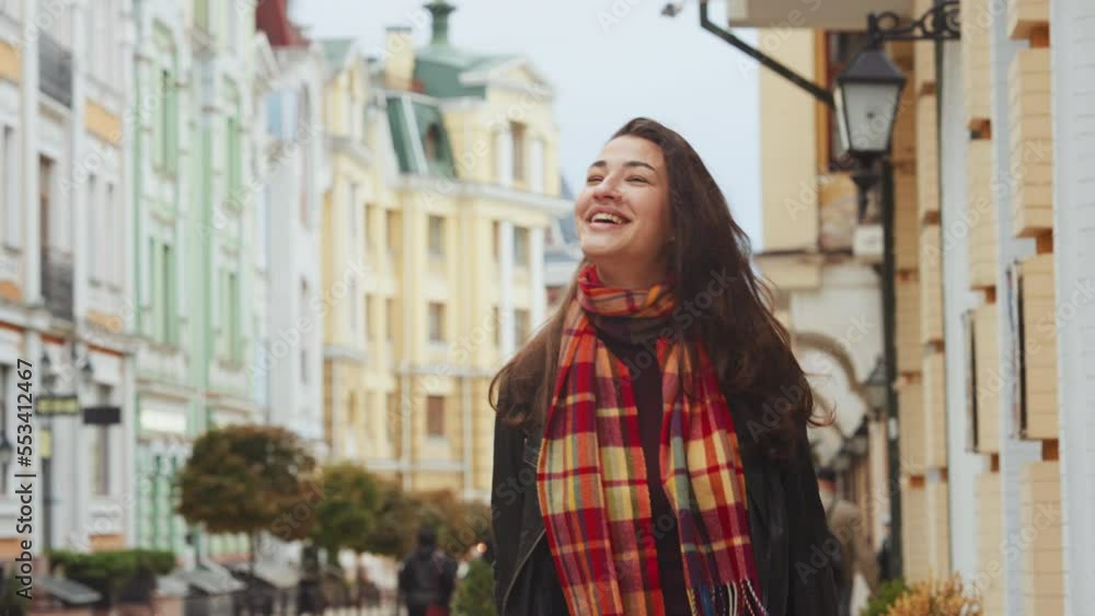 Young woman wearing leather jacket with checkered scarf walking in autumn city, looking at camera, blurred old buildings background. Tracking shot of happy person outside. Concept of joy Young woman wearing leather jacket with checkered scarf walking in autumn city, looking at camera, blurred old buildings background. Tracking shot of happy person outside. Concept of joy