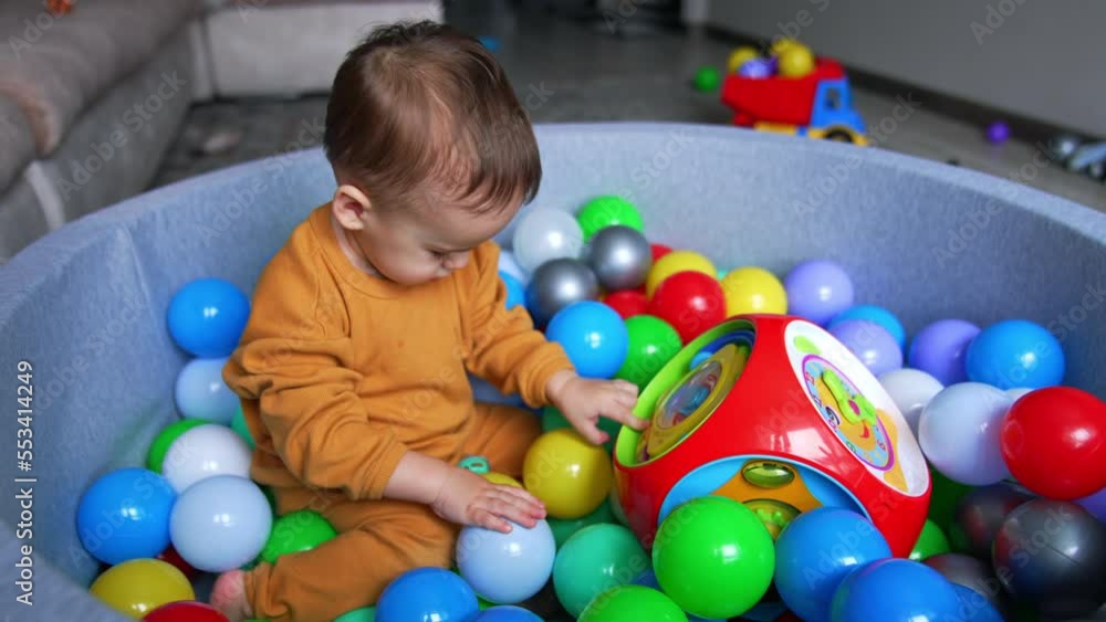 Calm toddler sitting in dry basin with balls. Beautiful baby touches the bright toy but then distracts on the pacifier. Calm toddler sitting in dry basin with balls. Beautiful baby touches the bright toy but then distracts on the pacifier.