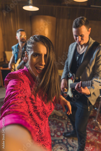 A caucasian woman singer in vivid pink jacket taking selfie with her music band. Vertical indoor shot. Blurred background. High quality photo