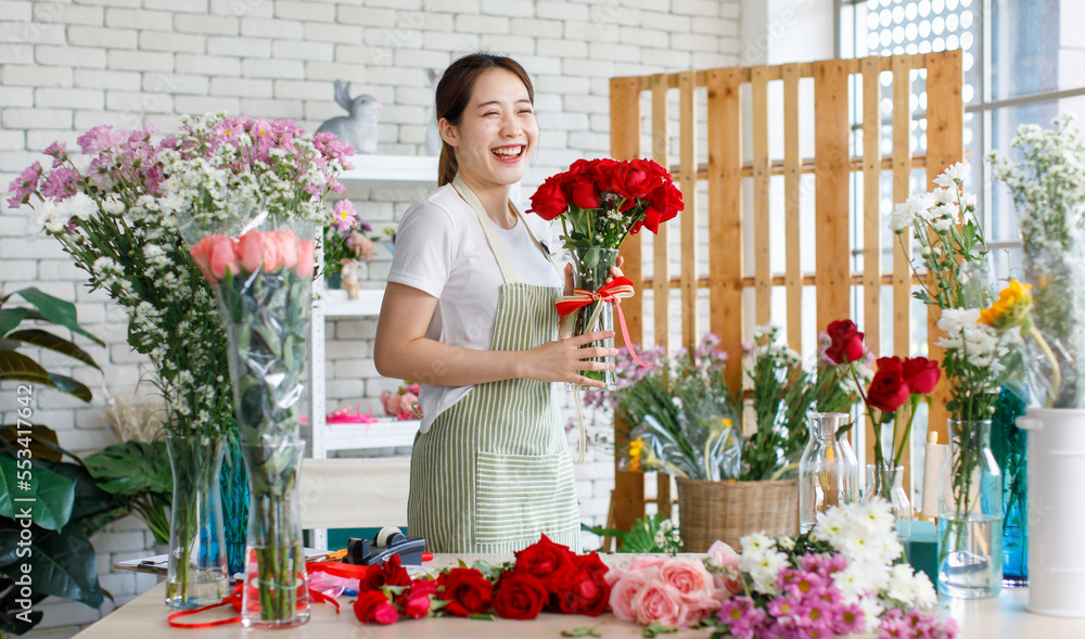 Millennial Asian young beautiful professional female flower shopkeeper owner decorator wearing apron standing smiling smelling decorating red roses bunch bouquet in glass vase with ribbon in store