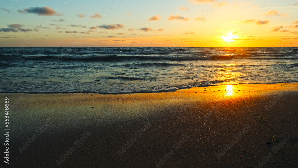 Beautiful sandy beach while sunset. Beautiful seascape of a sandy beach. Sun over horizon and  rolling ocean waves to the shore while sunset. Summer, travel and tourism. Beautiful wild nature.