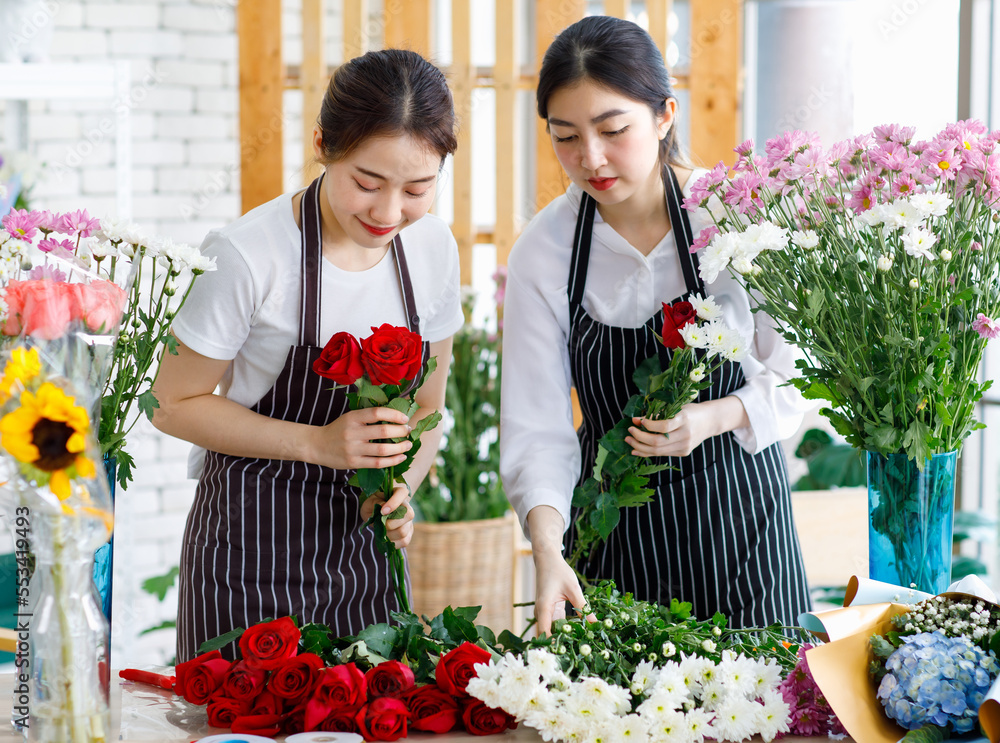 Millennial Asian young female flower shopkeeper decorator florist worker in apron smiling holding flower bunch bouquet colleague arranging decorating stalk in floral store.